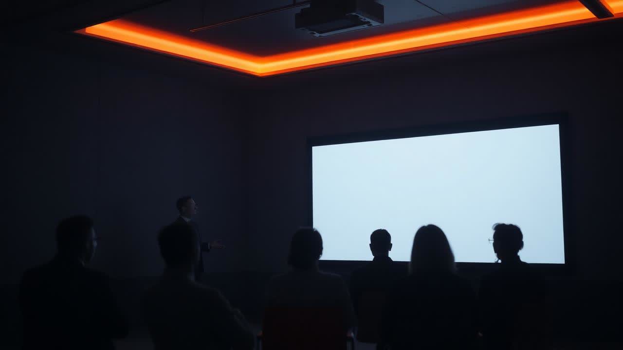 A presenter briefing a seated audience in a darkened room lit by a screen, with an orange ceiling light band